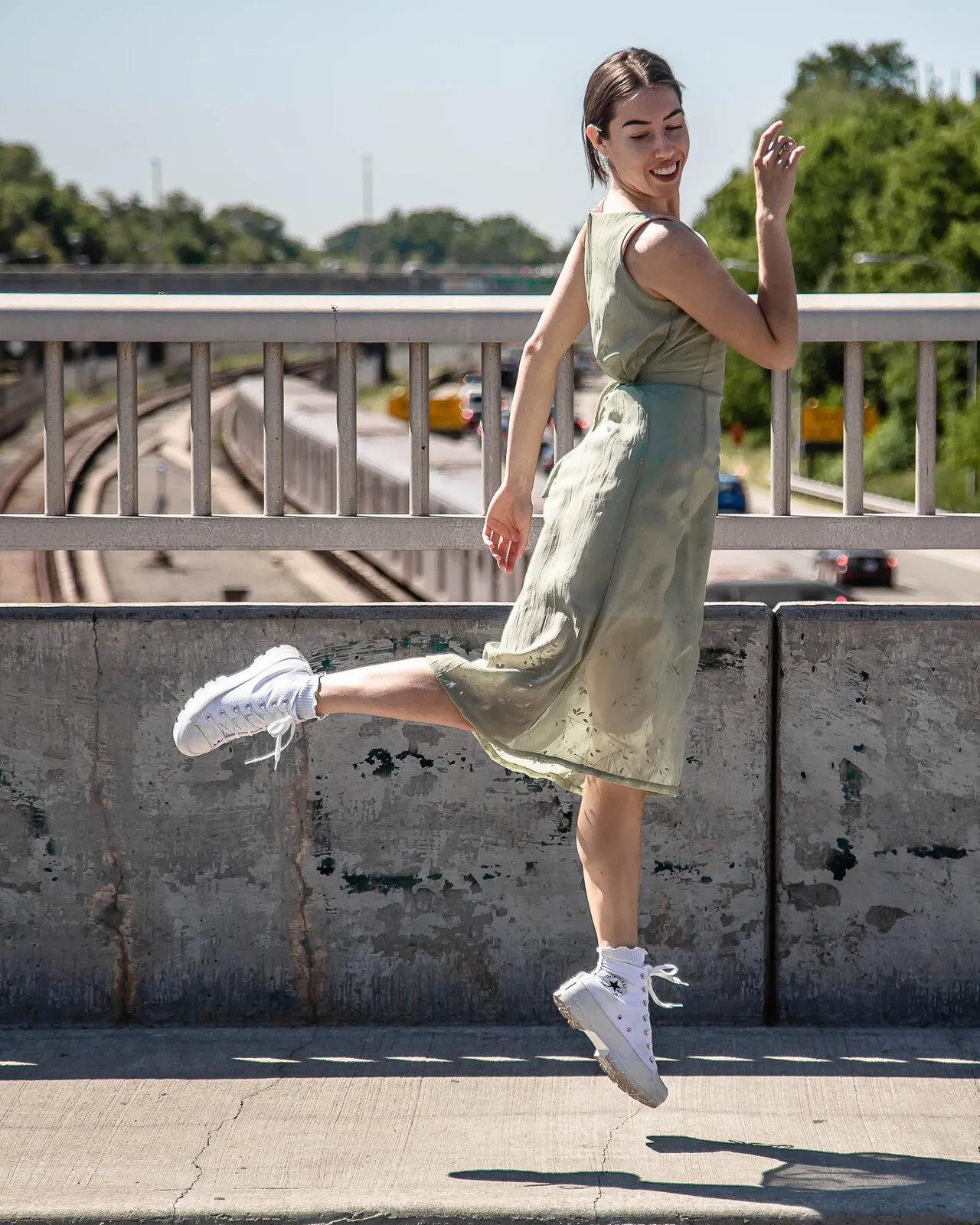 Mid-leap on a city bridge in a sage green dress and white sneakers, laughing